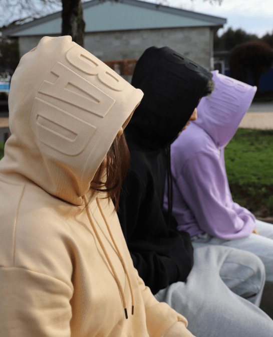 Models sitting side on wearing Due Hoodies in Tan, Black & Lavender